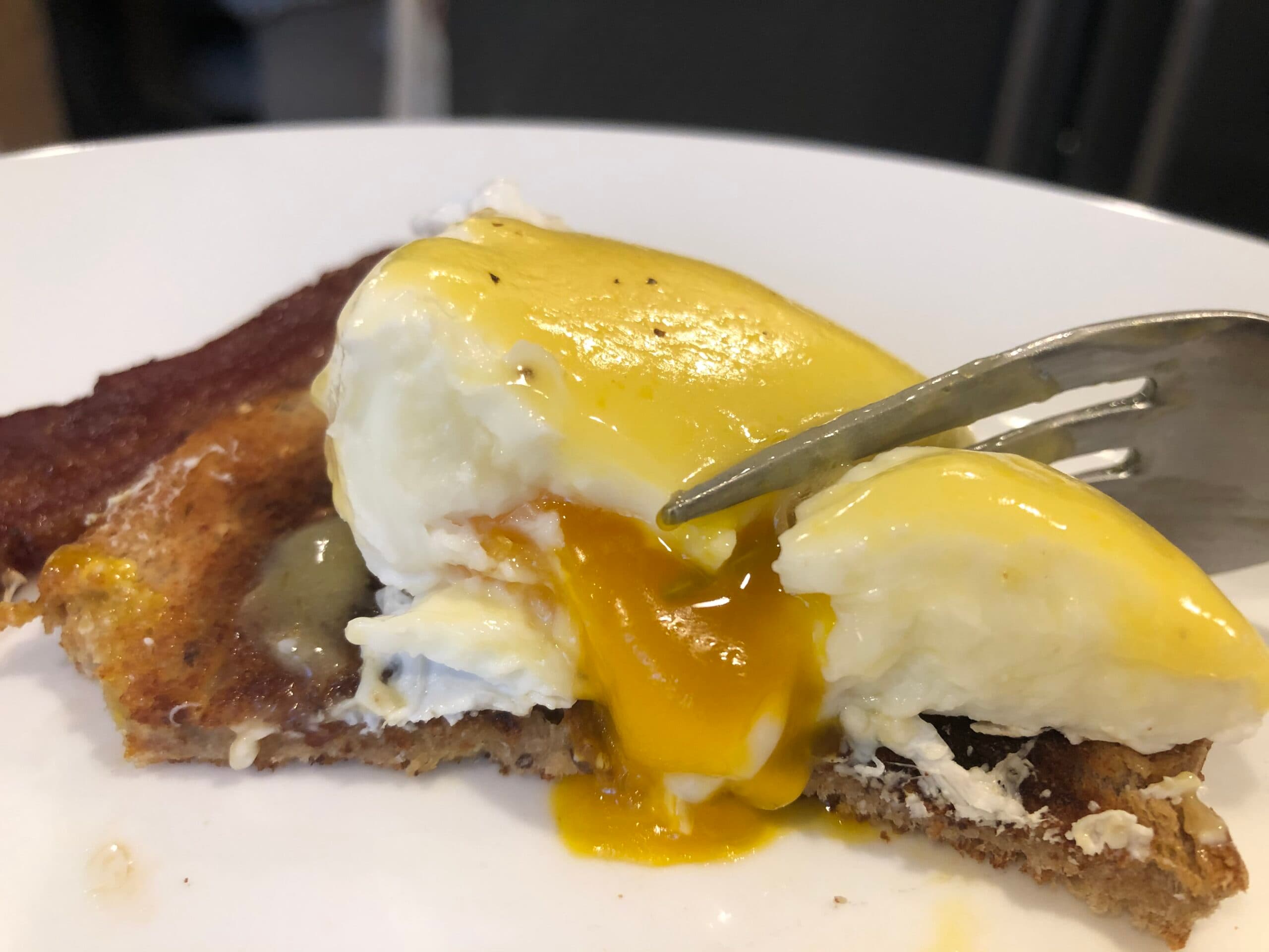 A close-up of a poached egg drizzled with hollandaise sauce on toasted bread, with a fork poised to cut into it.