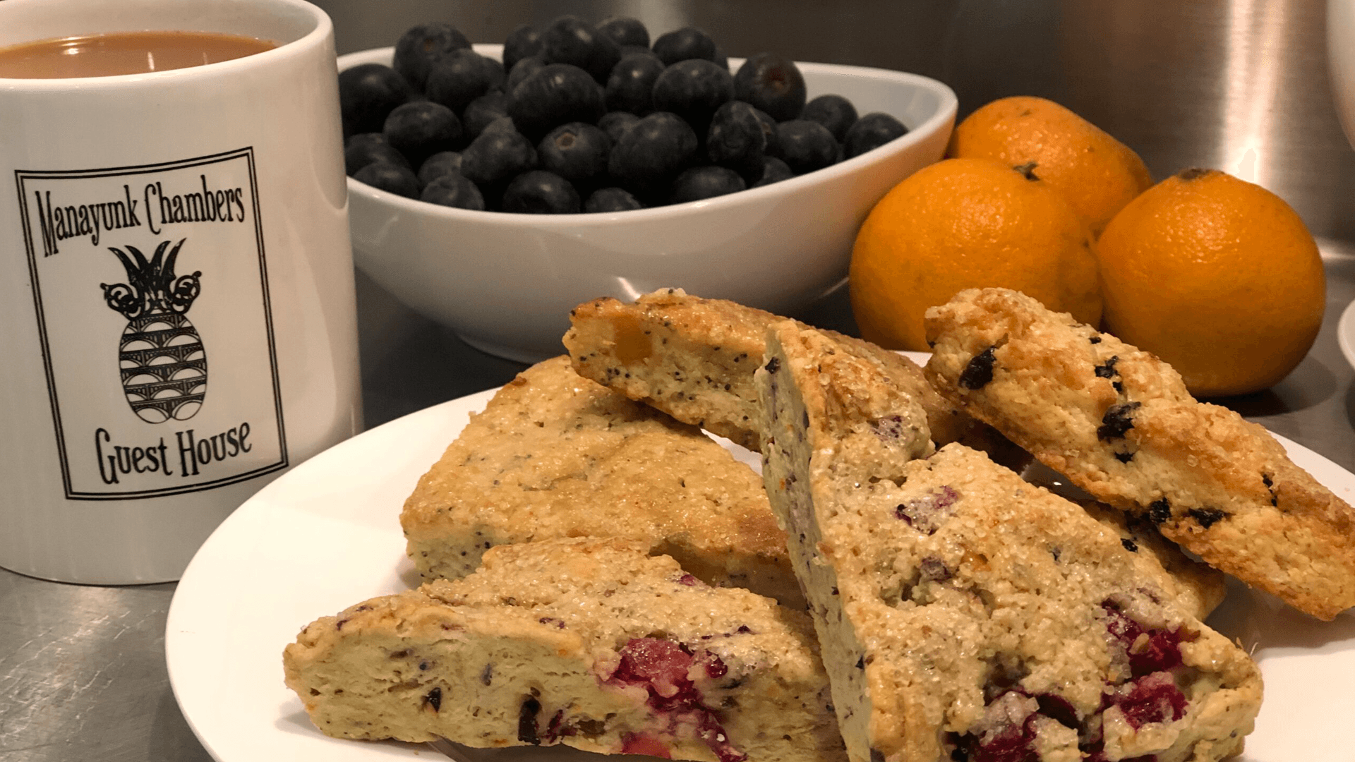 A cup of coffee, a bowl of blueberries, and a plate of scones next to oranges.