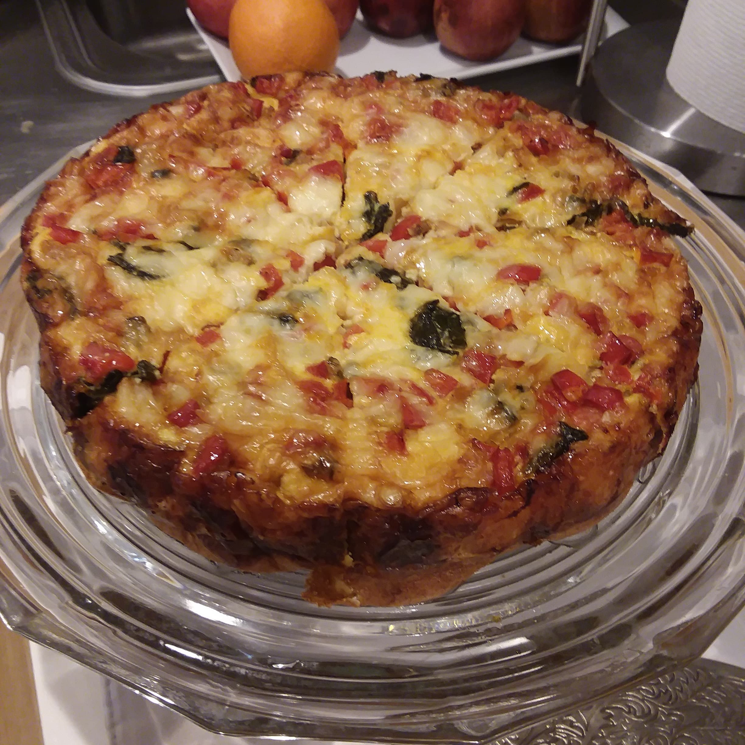A round, baked casserole topped with cheese and colorful vegetables, displayed on a glass platter.