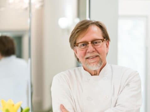 A smiling chef in a white coat stands confidently with arms crossed in a bright kitchen.