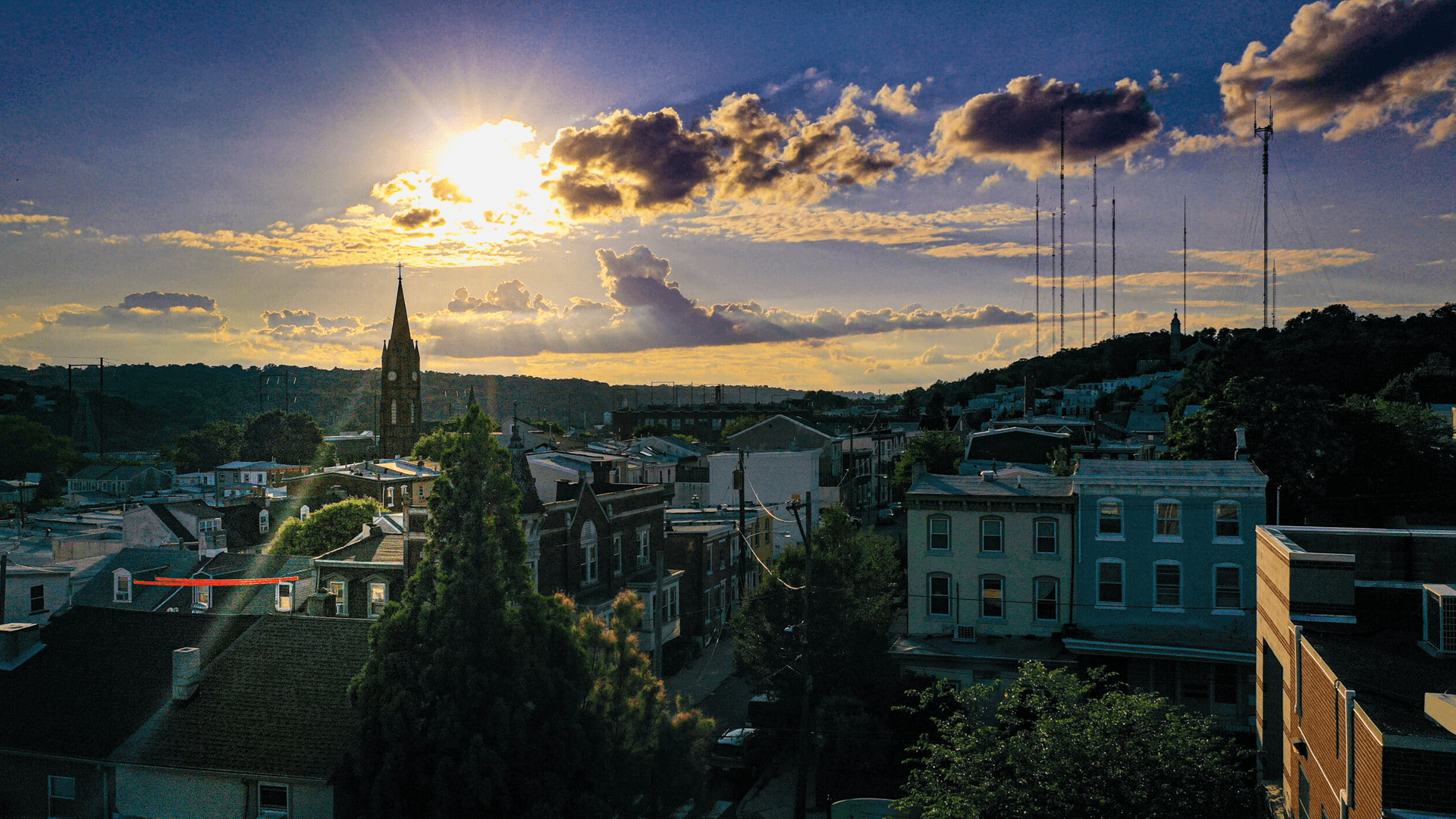 Sunset over a town featuring rooftops, a steeple, and communication towers.