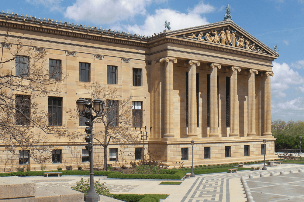 Facade of a classical building with columns, decorative sculptures, and surrounding landscaping.