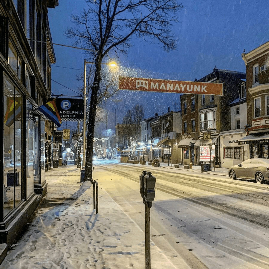 Snow-covered street in Manayunk during twilight, with shops lining the road and festive lights.